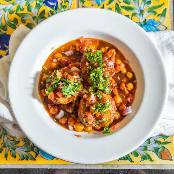 A white bowl of Indian aloo tikki chaat with chickpeas, sauce, onions, and cilantro.
