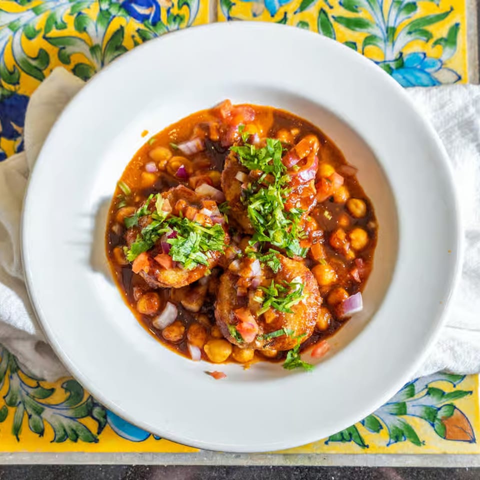 A white bowl of Indian aloo tikki chaat with chickpeas, sauce, onions, and cilantro.