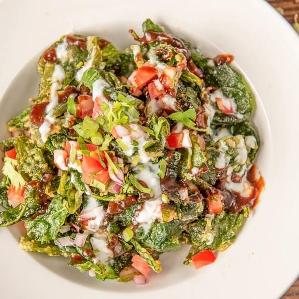 Crispy spinach chaat with yogurt, tamarind chutney, diced tomatoes, and onions in a white bowl.